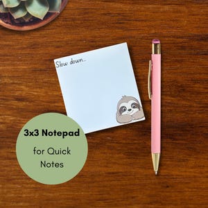 May include: A square white notepad with the text Slow down... and a cartoon sloth illustration in the bottom right corner, accompanied by a pink pen with gold accents. The notepad is labeled as a 3x3 notepad for quick notes.