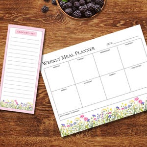 Peut inclure: Un planificateur de repas hebdomadaire et un bloc-notes de liste de courses avec un fond blanc, du texte noir et une bordure florale colorée en bas. Le planificateur comprend des sections pour chaque jour de la semaine et la mention Don't Forget To Defrost.