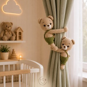 May include: Two beige crocheted teddy bears with dark brown embroidered noses and eyes, wearing green overalls with small buttons, are wrapped around a sage green curtain. The bears have round ears and are positioned one above the other.