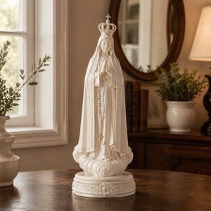 May include: A white statue of the Virgin Mary wearing a crown and a long, flowing robe with intricate detailing. The figure stands on a cloud-shaped base atop a circular pedestal, with hands clasped in prayer and a rosary hanging from the hands.