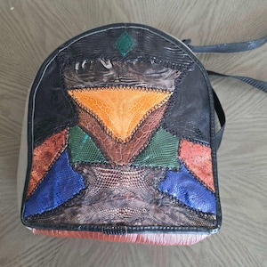 May include: A rounded backpack featuring a patchwork design of textured leather panels in black, orange, green, blue, and brown. The bag includes a small green diamond accent at the top, visible stitching between the panels, and black straps.