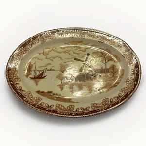 May include: An oval ceramic platter featuring a brown transferware print on a cream background. The central scene depicts a windmill, a sailboat on water, and trees. The wide rim is decorated with a repeating pattern of oak leaves and acorns.