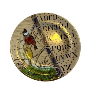 May include: A round ceramic plate featuring a split design. One side displays two birds perched on branches above green grass. The other side features a cream background with a repeating small pattern and the black capital letters A through Z.