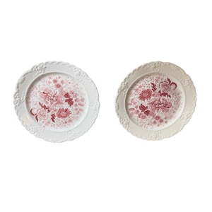 May include: Two round ceramic plates featuring a white base with a scalloped, embossed floral rim. The centre of each plate displays a detailed, intricate botanical pattern in a deep red colour, showcasing various flowers and foliage designs.