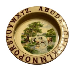 May include: A round, cream-colored ceramic plate featuring a dark red alphabet border from A to Z. The center displays a colorful pastoral scene of cows standing in a shallow stream near a large green tree under a light blue sky.