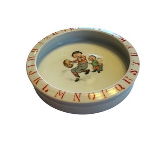 May include: A round white ceramic dish featuring a central illustration of a child holding a football with two other children nearby. The rim is decorated with the letters of the alphabet in red, starting with A and ending with Z in a circular pattern.