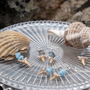 May include: A jewellery set featuring two necklaces and two pairs of earrings. Each piece incorporates a small, tan shark tooth wrapped in silver wire, accented with either light blue or grey stone beads on thin silver-toned metal chains.