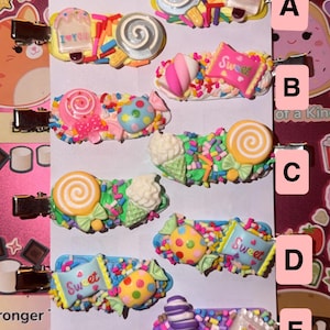 May include: A collection of ten hair clips featuring colorful miniature candy and dessert decorations, including lollipops, ice cream, and candy bags with the text Sweet, arranged in pairs labeled A through E on a white rectangular card.