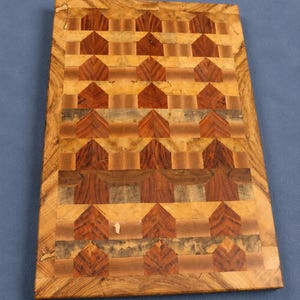 Könnte beinhalten: Ein rechteckiges Schneidebrett aus Holz mit einem komplexen Hirnholz-Mosaikmuster. Das Design besteht aus sich wiederholenden hausförmigen Motiven in verschiedenen Braun-, Beige- und Grautönen, umrahmt von einem hellbraunen Holzrand.