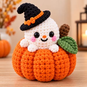 Könnte beinhalten: Eine gehäkelte Amigurumi-Figur, bestehend aus einem weißen Geist mit schwarzen Augen und rosa Wangen, der einen schwarzen Hexenhut mit orangefarbenem Band trägt und in einem strukturierten orangefarbenen Kürbis mit braunem Stiel und grünem Blatt sitzt.