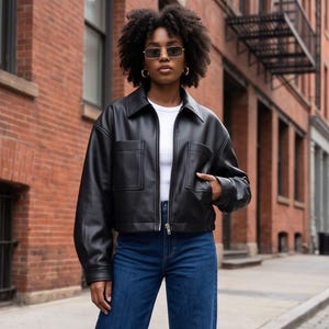 Puede incluir: Una chaqueta de cuero negra con cuello, cierre de cremallera frontal y dos bolsillos grandes en el pecho. La chaqueta se lleva sobre una camiseta blanca lisa y se combina con vaqueros azules de talle alto. El conjunto incluye gafas de sol rectangulares.