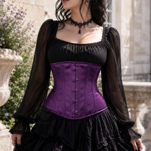 May include: A deep purple satin corset featuring a front busk closure with silver-toned metal clasps. The corset is worn over a black long-sleeved top with sheer sleeves and a black lace choker necklace with a dark teardrop pendant.