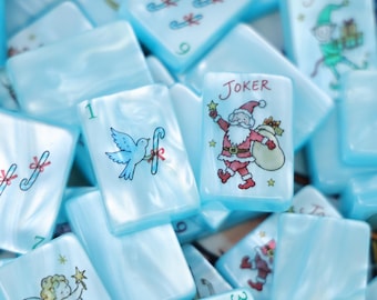 Könnte beinhalten: Eine Sammlung hellblauer, perlmuttartiger Mahjong-Spielsteine mit weihnachtlichen Illustrationen. Die Steine zeigen verschiedene Motive, darunter den Weihnachtsmann mit dem Schriftzug JOKER, eine Taube mit Zuckerstange, einen Engel und Zuckerstangen.