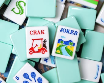 Könnte beinhalten: Eine Ansammlung rechteckiger Mahjong-Spielsteine mit hellminzgrüner Rückseite und weißer Vorderseite. Die Steine zeigen verschiedene bunte Illustrationen, darunter ein rotes Golfcart mit der Aufschrift CRAK, ein JOKER-Stein mit Landschaft.