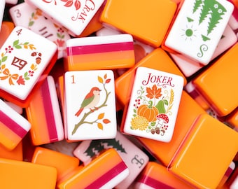 Könnte beinhalten: Eine Sammlung rechteckiger Mahjong-Spielsteine mit herbstlichen Motiven. Die Steine zeigen Illustrationen von Kürbissen, Vögeln, Äpfeln und Laub in den Farben Orange, Rot und Grün. Einige Steine tragen den Text JOKER oder CRAK.