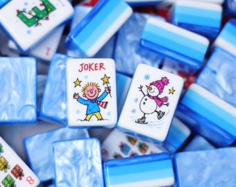May include: A collection of rectangular mahjong tiles featuring a blue and white marbled pattern. Several tiles display colorful illustrations of gift boxes, a snowman, and a character labeled JOKER with a star wand, alongside numerical markings.