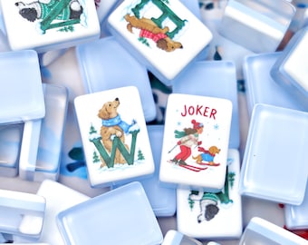 Könnte beinhalten: Eine Sammlung rechteckiger Mahjong-Spielsteine mit hellblauer Rückseite und weißer Vorderseite. Die Steine zeigen bunte Illustrationen von Hunden in Winterkleidung, verschneite Landschaften und das Wort JOKER in roter Schrift.
