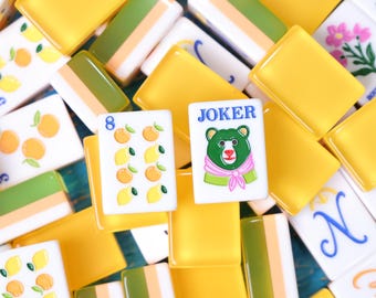 May include: A collection of mahjong tiles featuring yellow and green backs, with white faces displaying various illustrations including lemons, oranges, flowers, and a Joker tile with a bear wearing a pink scarf and the text JOKER printed in blue.