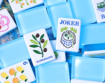 Könnte beinhalten: Eine Sammlung rechteckiger Mahjong-Spielsteine mit einer durchscheinenden hellblauen Basis und weißen Oberseiten. Die Steine zeigen verschiedene bunte Illustrationen, darunter eine Eule mit dem Text JOKER, einen Olivenzweig mit der Aufschrift OLIBEBRANCH, Früchte, ein Boot und einen Hund.