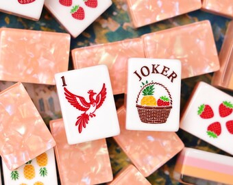 Könnte beinhalten: Eine Sammlung von Mahjong-Steinen, bestehend aus weißen Steinen mit roten Erdbeer- und gelben Ananas-Illustrationen, einem Stein mit einem roten Phönix und der Zahl 1 sowie einem Stein mit einem braunen Obstkorb und dem Text JOKER.