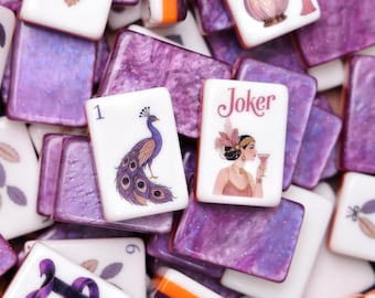 Könnte beinhalten: Eine Sammlung rechteckiger Mahjong-Spielsteine mit violetter Marmorierung. Die Spielsteine zeigen verschiedene Illustrationen, darunter einen Pfau, eine Frau mit Glas und dem Text Joker, einen Parfümflakon und einen roten Edelstein.
