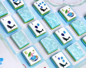 Könnte beinhalten: Ein Set rechteckiger Mahjong-Spielsteine mit weißer Vorderseite, blauen Wal-Illustrationen, Ziffern und grünen Meerjungfrauen-Grafiken. Einige Steine haben eine marmorierte hellblaue Rückseite, andere zeigen die Zahlen 1, 2, 4, 6, 9 oder den Buchstaben E.