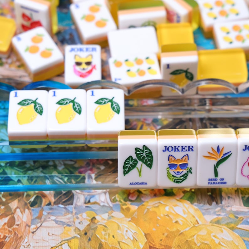 May include: A collection of white mahjong tiles with yellow backs, featuring various illustrations including lemons, oranges, Alocasia leaves, a Bird of Paradise flower, and a Joker character wearing sunglasses, arranged on a clear tiered display rack.