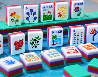 Könnte beinhalten: Eine Sammlung rechteckiger Mahjong-Spielsteine mit farbenfrohen Illustrationen wie Blumen, Tennisschlägern, Golffahnen, Palmen und dem Wort JOKER. Jeder Spielstein hat eine weiße Vorderseite und einen mehrschichtigen Sockel in Grün, Rot und Lila.