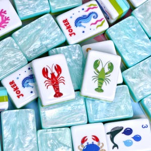 May include: A collection of rectangular mahjong tiles featuring a marbled light blue and white finish. The tiles display various colourful illustrations including a red lobster, a green lobster, a blue seahorse with the text Joker, and a blue crab.