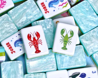 May include: A collection of rectangular mahjong tiles featuring a marbled light blue and white finish. The tiles display various colorful illustrations including a red lobster, a green lobster, a blue seahorse with the text Joker, and a blue crab.