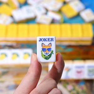 May include: A white rectangular mahjong tile featuring the word JOKER in blue text above a colorful illustration of a fox wearing blue sunglasses and a green scarf, with other blurred yellow and white mahjong tiles visible in the background.