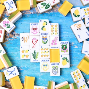May include: A collection of rectangular mahjong tiles with white faces and yellow backs. The tiles feature colorful illustrations of lemons, flowers, rosemary, and animals, including a poodle, fox, bear, and owl, with the word JOKER printed on some.