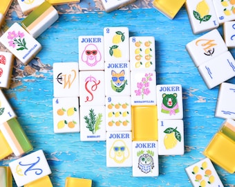 Könnte beinhalten: Eine Sammlung rechteckiger Mahjong-Spielsteine mit weißen Vorderseiten und gelben Rückseiten. Die Steine zeigen bunte Illustrationen von Zitronen, Blumen, Rosmarin und Tieren, darunter ein Pudel, ein Fuchs, ein Bär und eine Eule mit dem Wort JOKER.