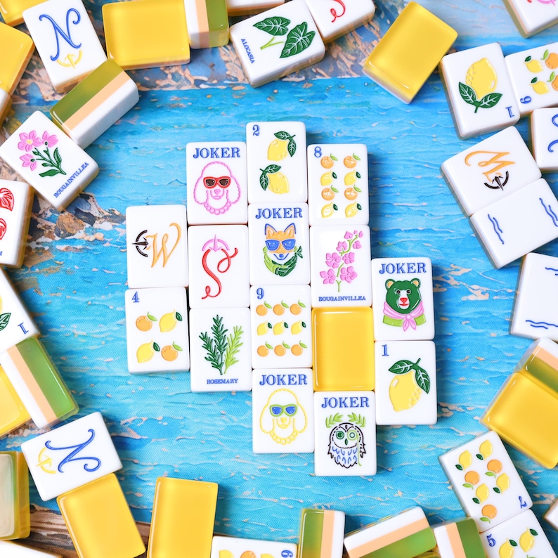 May include: A collection of rectangular mahjong tiles with white faces and yellow backs. The tiles feature colorful illustrations of lemons, flowers, rosemary, and animals, including a poodle, fox, bear, and owl, with the word JOKER printed on some.