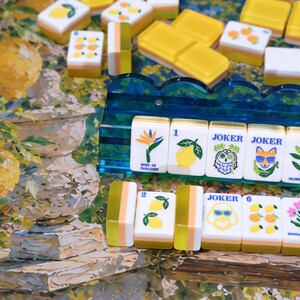 May include: A set of rectangular mahjong tiles featuring colorful illustrations of lemons, oranges, flowers, and animals, including a bird of paradise, an owl, and a fox wearing sunglasses, displayed on a blue acrylic rack and a patterned surface.