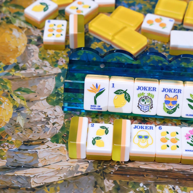 May include: A set of rectangular mahjong tiles featuring colorful illustrations of lemons, oranges, flowers, and animals, including a bird of paradise, an owl, and a fox wearing sunglasses, displayed on a blue acrylic rack and a patterned surface.