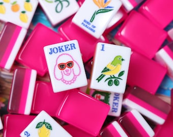 Könnte beinhalten: Eine Sammlung rechteckiger Mahjong-Spielsteine mit weißen Vorderseiten und leuchtend pinkfarbenen Rückseiten. Die Steine zeigen bunte Illustrationen, darunter einen Pudel mit Sonnenbrille und der Aufschrift JOKER, eine Paradiesvogelblume, einen gelben Vogel und Zitronen.