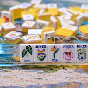 May include: A set of white mahjong tiles with yellow backs, featuring colorful illustrations and text. Five tiles in the foreground display an Alocasia plant, a fox wearing sunglasses, a bird of paradise flower, a poodle, and an owl with a monocle.