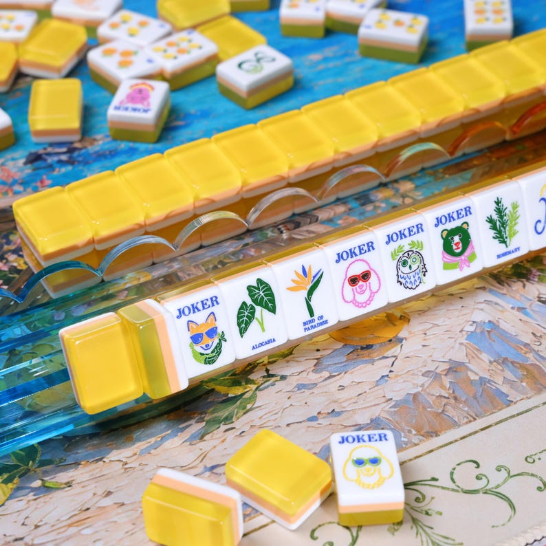 May include: A set of Mahjong tiles featuring translucent yellow backs and white fronts with various colorful illustrations, including animals wearing sunglasses, plants, and the text JOKER, ALOCASIA, BIRD OF PARADISE, and ROSEMARY, arranged on a blue tray.