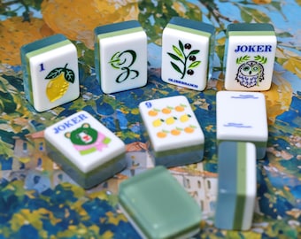 Könnte beinhalten: Eine Sammlung rechteckiger Mahjong-Spielsteine mit weißen Vorderseiten und grünen Rückseiten. Die Steine zeigen verschiedene aufgedruckte Motive, darunter eine Zitrone, die Zahl 3, einen Olivenzweig, eine Joker-Eule, einen Joker-Bären, die Zahl 9 mit Orangen.