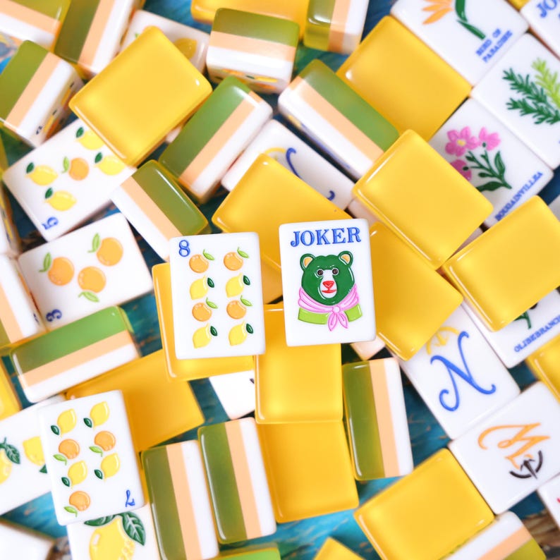May include: A collection of mahjong tiles featuring yellow and green backs, with white faces displaying various illustrations including lemons, oranges, flowers, and a Joker tile with a bear wearing a pink scarf and the text JOKER printed in blue.