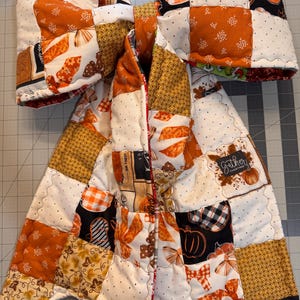 May include: A large, quilted fabric bow featuring a patchwork design of orange, white, and black patterns, including pumpkins, leaves, and plaid. A small black fabric tag on the lower right side displays the white text "gather together".