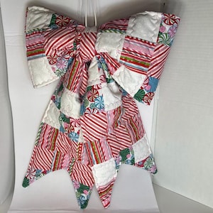 May include: A large, quilted fabric bow featuring a patchwork design of red, white, and green candy cane stripes, peppermint swirl patterns, and solid white squares with a subtle snowflake texture. The bow has two long, pointed tails.