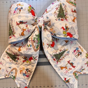 May include: A large, quilted fabric bow featuring a repeating pattern of characters from How the Grinch Stole Christmas, including the Grinch, Max the dog, and Cindy Lou Who, set against a white background with blue snowflakes and Christmas trees.