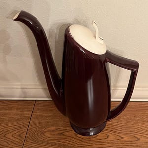 May include: A dark burgundy ceramic coffee pot with a white lid and a long, curved spout. The pot has a modern, mid-century design.