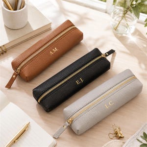 May include: Three rectangular pencil cases in tan, black, and light gray with a pebbled texture. Each case features a gold-toned metal zipper and is personalized with gold-embossed initials: A.M. on the tan, E.J. on the black, and L.C. on the gray.