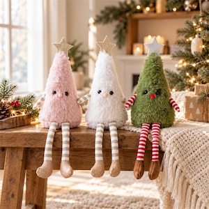 Puede incluir: Tres figuras de árbol de Navidad de peluche en forma de cono con texturas suaves en rosa, blanco y verde. Cada figura presenta una estrella dorada en la punta, ojos oscuros pequeños, una nariz redonda y piernas colgantes de tela a rayas.