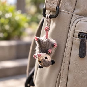 Puede incluir: Un pequeño llavero de zarigüeya tejido a ganchillo con un cuerpo gris difuso, cara beige claro, orejas negras y patas rosas. La zarigüeya cuelga boca abajo por su cola de un bolso de tela beige, sujeta con un pequeño lazo de cuerda anudado.