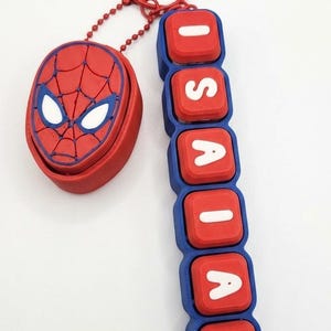 May include: A red and blue keychain featuring a circular charm with a spider web pattern and white eyes, attached to a vertical row of six red square blocks with white letters spelling ISAIAH, all connected by a red ball chain ring.