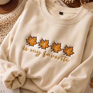 Op de afbeelding: Een crèmekleurige sweater met ronde hals en een geborduurd ontwerp van vier oranje esdoornbladeren. Elk blad bevat een letter die het woord FALL vormt. Onder de bladeren is de tekst is my favorite in een cursief lettertype geborduurd.